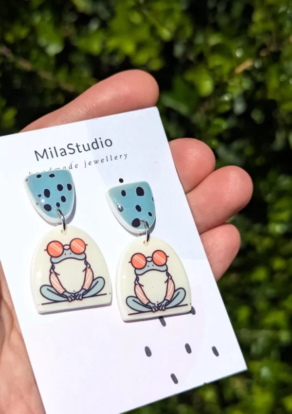 A hand holds a pair of frog-themed earrings, showcasing their playful design and vibrant colors.
